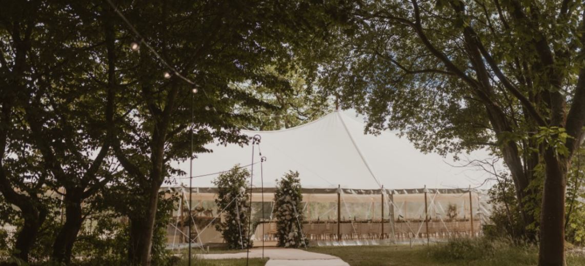 A large white event tent is set up outdoors, surrounded by trees and greenery. There are string lights hanging and long tables arranged inside the tent, suggesting a formal gathering or celebration.